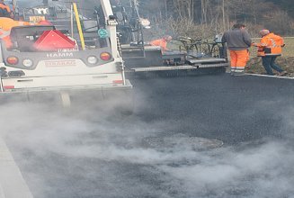Ein Fertiger von Hinten mit dampfenden Asphalt auf dem die Bauarbeitenden die Maschine steuern und den Asphalt prüfen.
