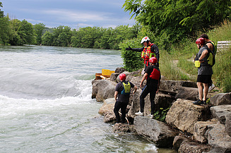 Teilnehmende eines Wasserrettungskurses stehen in Schutzkleidung auf Felsen am Flussufer und beobachten die Strömung des Wassers.