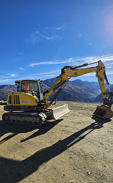 Gelber STRABAG Bagger auf einer geraden Sandfläche. Im Hintergrund blauer Himmel mit strahlendem Sonnenschein und einem Bergpanorama