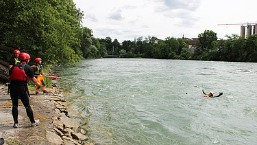 Teilnehmende eines Wasserrettungskurses stehen in Schutzkleidung am Ufer eines Flusses, während eine Person im Wasser eine Rettungsübung in der Strömung durchführt.