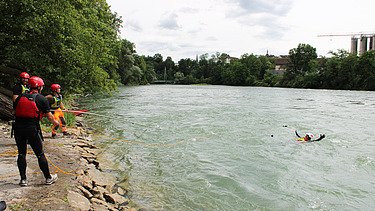 Teilnehmende eines Wasserrettungskurses stehen in Schutzkleidung am Ufer eines Flusses, während eine Person im Wasser eine Rettungsübung in der Strömung durchführt.
