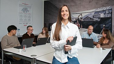 Kalkulatorin Katarina steht m Büro. Im Hintergund sind Kolleg:innen zu sehen.