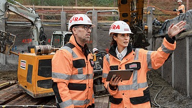 Frau mit Helm und Baustellenkleidung erklärt Mann die Baustelle
