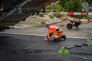 Ein Bauarbeiter verteilt heissen Asphalt mit einer Schaufel neben einer Strassenbaumaschine.