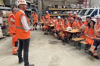 Lernende in orangefarbener STRABAG Arbeitskleidung sitzen auf Festbänken und hören zwei Personen zu, die vor ihnen sprechen
