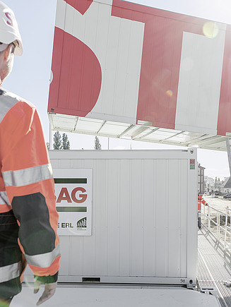 STRABAG - Mitarbeiter in oranger Schutzkleidung beobachten das Absetzen eines grossen Containers mit STRABAG-Aufschrift durch einen Kran auf einer Baustelle.