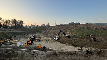 Mehrere Baumaschinen und Lastwagen arbeiten auf einer grossen Erdbaustelle im Burgaurfeld, im Hintergrund sind Hänge und Bäume zu sehen.