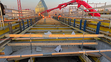  Ein Kran der STRABAG hebt nachts ein langes Brückenteil über Bahngleise. Die Baustelle ist hell erleuchtet.