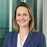 Kaufmännische Bereichsleiterin im Anzug lächelt in die Kamera. Im Hintergrund ist ein Lift zu sehen.