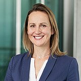 Kaufmännische Bereichsleiterin im Blazer lächelt in die Kamera. Im Hintergrund ist ein Lift zu sehen.