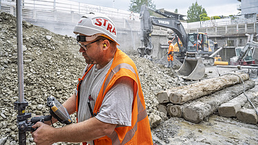 Der Polier in der Baugrube beim Vermessen am Lasermessgerät im Hintergrund sieht man das restliche Baustellenpersonal und zwei Bagger