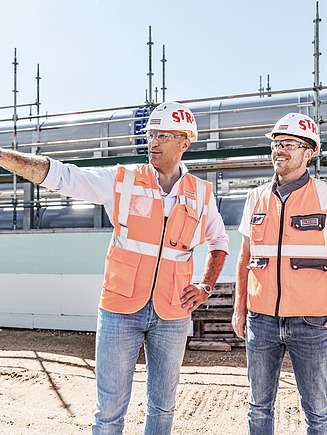 Drei STRABAG - Mitarbeiter mit Helmen und Warnwesten stehen auf einer Baustelle, einer zeigt in die Ferne und bespricht den Baufortschritt.