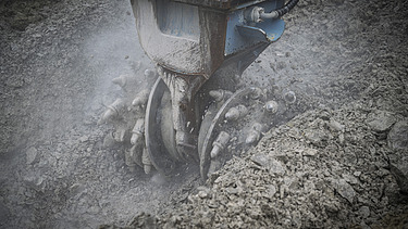 Ein Bagger trägt den Boden ab mit einem Fräserbohrkopf, es staubt 