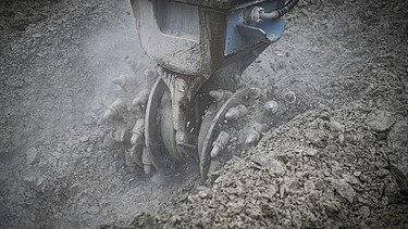 Ein Bagger trägt den Boden ab mit einem Fräserbohrkopf, es staubt 
