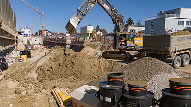 Bagger hebt Loch aus. Daneben ein LKW wo den Aushub abtransportiert