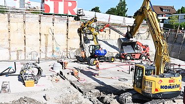 Mehrere Baufahrzeuge in einer Baugrube bei der Arbeit. Im Hintergrund ein weisser STRABAG Container mit der Aufschrift STR in grossen roten Buchstaben.