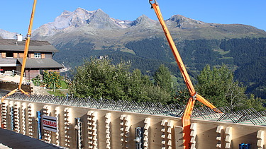 Ein langer Holzträger hängt an den Seilen eines Krans und wird über der Baustelle vor einer Berglandschaft positioniert.