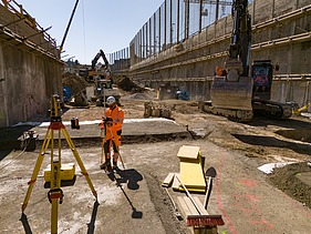 Rüschlikon Bahnhof Süd – STRABAG Tiefbau und Spezialtiefbau Vermesser steht in der Baugrube am Ausmessen. Rechts daneben hebt ein Bagger gerade ein Loch aus.