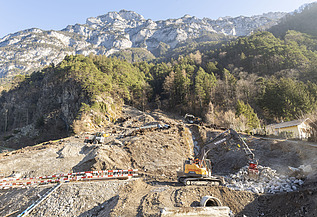 Ein Bagger der STRABAG räumt Geröll an einem steilen Hang unterhalb einer Felswand weg. Baustelle zur Hangsicherung im Gebirge.