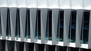 Blick von vorne auf die markante Fassade: Die Glasflächen sind durch dreieckig zulaufende, vertikal angeordnete Fassadenelemente rhythmisch strukturiert. Die Geometrie erzeugt ein prägnantes Licht- und Schattenspiel. 