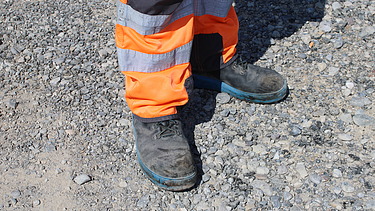 Bauarbeiter in orangefarbener Warnkleidung steht mit robusten Arbeitsschuhen auf einer kiesigen Fläche.