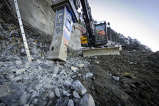 Bagger spitzt Felsgestein an einer Baugrube auf; im Vordergrund liegen aufgebrochene Geröllstücke.