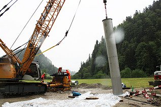 Baustellenkran hebt Filterbrunnen hoch, während STRABAG Bauarbeiter am Ziel warten. Im Hintergrund sind grüne, bewaldete Berge zu sehen.