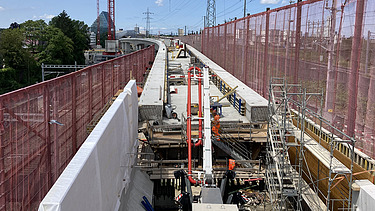  Ein Kran der STRABAG hebt nachts ein langes Brückenteil über Bahngleise. Die Baustelle ist hell erleuchtet.