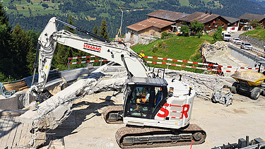Weisser STRABAG-Abbruchbager bricht Teile der ehemaligen Lochlitobelbrücke ab. Im Hintergrund Schweizer Berge.