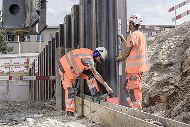 Zwei Bauarbeiter in orangefarbener Schutzkleidung befestigen grosse Stahlplatten im Boden. Die Platten bilden eine Spundwand.