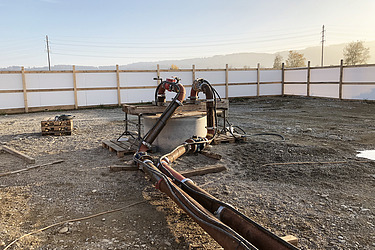 Auf einer Baustelle sind mehrere dicke Rohre und Schläuche verlegt, die zu einer Pumpe führen. Die Anlage dient zur Grundwasserabsenkung, der Boden ist feucht und von einem Bauzaun umgeben.