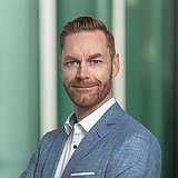 Kaufmännischer Bereichsleiter im Anzug lächelt in die Kamera. Im Hintergrund ist ein Lift zu sehen.