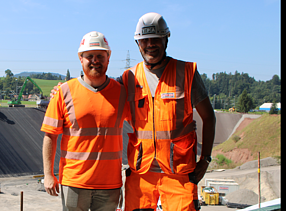 Zwei Männer in orangefarbener Bauausrüstung stehen lächelnd nebeneinander auf einer Baustelle mit Erdwall im Hintergrund.