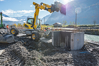 Ein orangener Bagger arbeitet auf einer Baustelle vor einer alpinen Bergkulisse. Neben dem Bagger steht ein grosser, runder Betonfundamentkörper, umgeben von Schotter und Erde. 