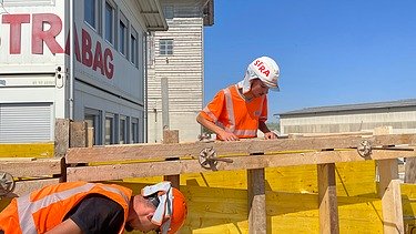 Zwei Lernende in orangefarbener STRABAG Arbeitskleidung arbeiten an einer gelben Verschalung auf einer Baustelle.