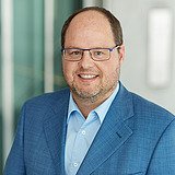 Technischer Bereichsleiter im Anzug lächelt in die Kamera. Im Hintergrund ist ein Lift zu sehen.