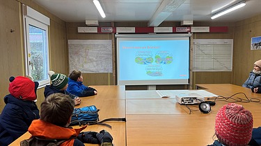 Eine Schulklasse sitzt in einem Baucontainer und verfolgt eine Präsentation zum Brückenbau auf einer Leinwand.