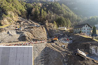 STRABAG-Baustelle am Hang: Volvo-Bagger und Dumper bei Erdarbeiten neben einer grossen Betonmauer in bergiger Umgebung.