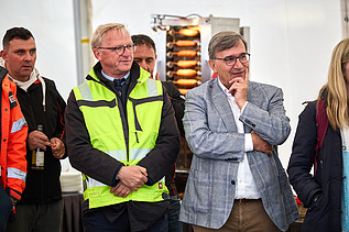 Mehrere Personen stehen bei einer Veranstaltung zusammen. Eine Person in einer gelben Sicherheitsjacke und eine andere im grauen Sakko stehen im Vordergrund und hören aufmerksam zu.