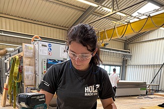 Holzbauhalle im Vordergrund die Zimmerin on Tour mit einem Herz aus Holz in der Hand. Im Hintergrund die CNC-Maschine