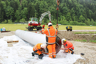 Drei STRABAG Bauarbeiter in orangefarbener Arbeitskleidung bereiten den Filterbrunnen für das Einsetzen mit einem Kran vor; im Hintergrund ein Bagger und Bäume.