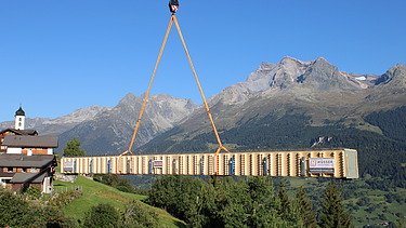 Ein grosser Pneukran hebt einen langen Holzträger hoch in die Luft im Hintergrund ist eine Berglandschaft und ein Dorf zu sehen.