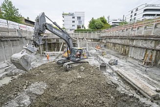 Ein Bagger steht auf Aushub in einer Baugrube hebt gerade die Schaufel an, im Hintergrund ist das Baustellenpersonal zu sehen