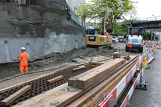Baustelle an einer Strasse mit Nagelwand, mehreren Baumaschinen und einem Arbeiter in oranger Kleidung an einem bewölkten Tag.