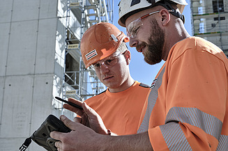 Zwei STRABAG - Mitarbeiter aus dem Hochbau in orangefarbener Arbeitskleidung und Helmen betrachten ein Tablet auf einer Baustelle.