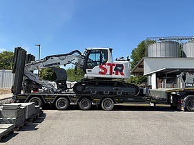 Anlieferung vom weisser STRABAG Liebherr-Raupenbagger, dieser steht verladen auf einem Tieflader-LKW