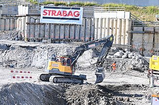  Ein gelber Bagger in einer Baugrube bei der Arbeit. Im Hintergrund sieht man ein STRABAG Banner 