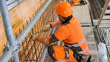  Zwei Bauarbeiter in orangefarbener Warnkleidung von STRABAG arbeiten auf einem Gerüst an einer Bewehrung.