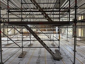 Innenaufnahme vom Landhaus mit einem umfangreichen Gerüstsystem und einer zentralen Treppe aus Metall. Der Boden ist mit Schutzfolie abgedeckt, auf der „MAXIMUM“ aufgedruckt ist. Das Gerüst reicht bis zur Decke und ermöglicht Arbeiten auf mehreren Ebenen im grossen, lichtdurchfluteten Raum mit hohen Fenstern.
