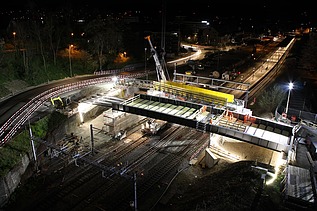 Baustelle in Küssnacht bei Nacht, beleuchtet von Scheinwerfern. Über den Bahngleisen wird eine neue Bahnbrücke mit einem grossen Kran eingesetzt. Im Hintergrund sind ein Bahnhof, Strassen mit Lichtspuren und beleuchtete Gebäude zu sehen.